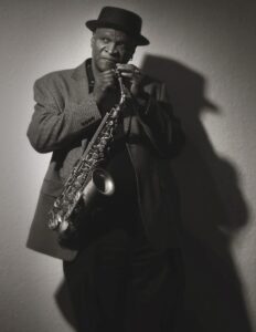 A black and white picture of an African American man posing in a black hat, holding a saxophone