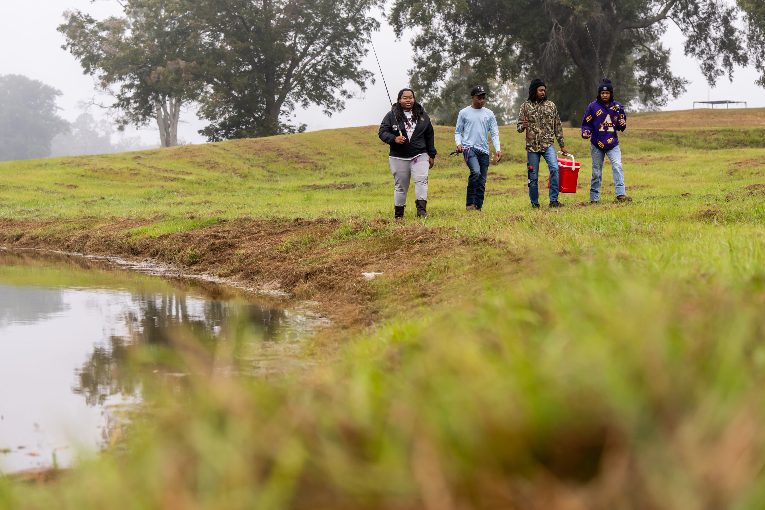 Four Alcorn State University students walking along the edge of a pond carrying fishing equipment.
