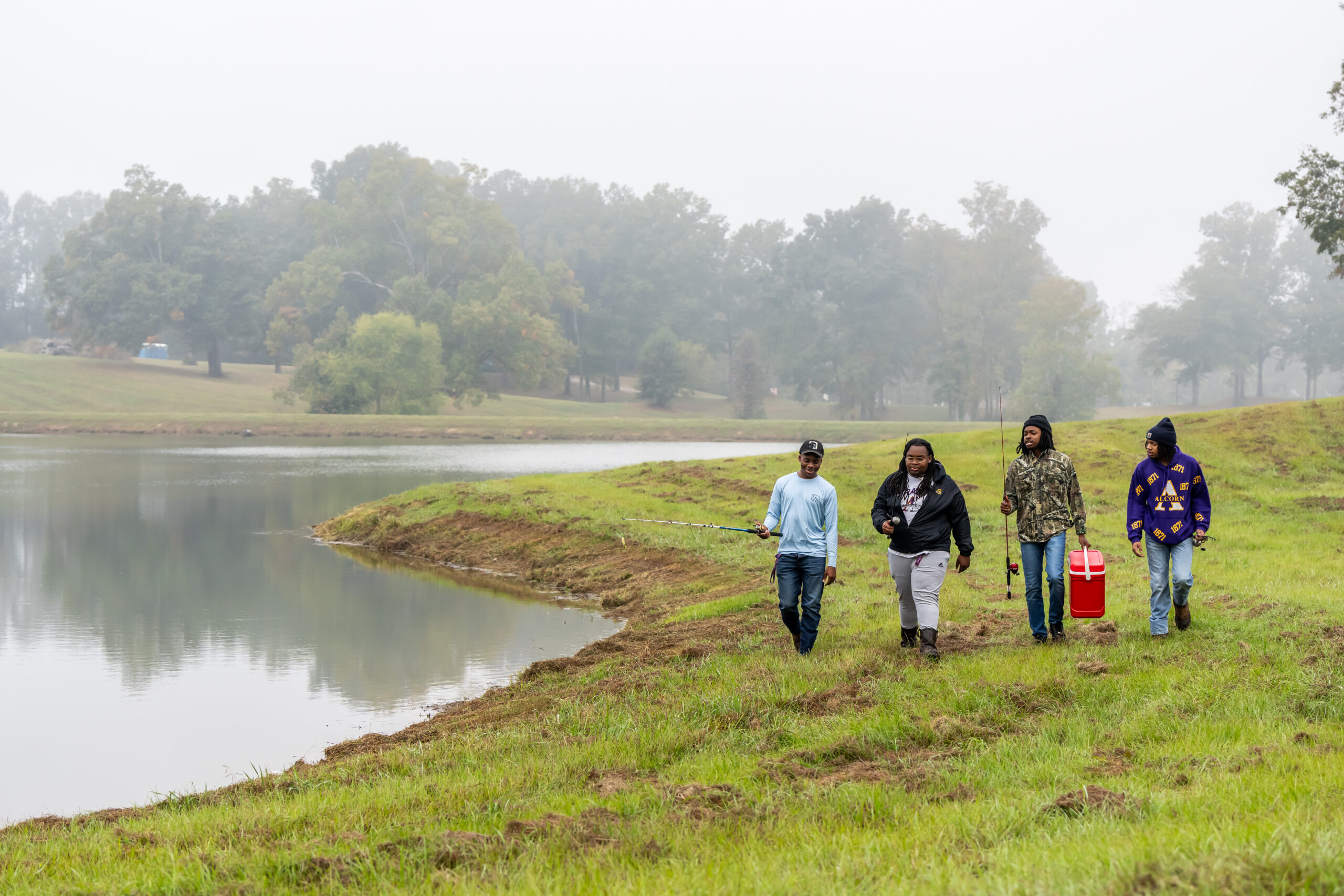 Four Alcorn State University students walking along the edge of a pond carrying fishing equipment.