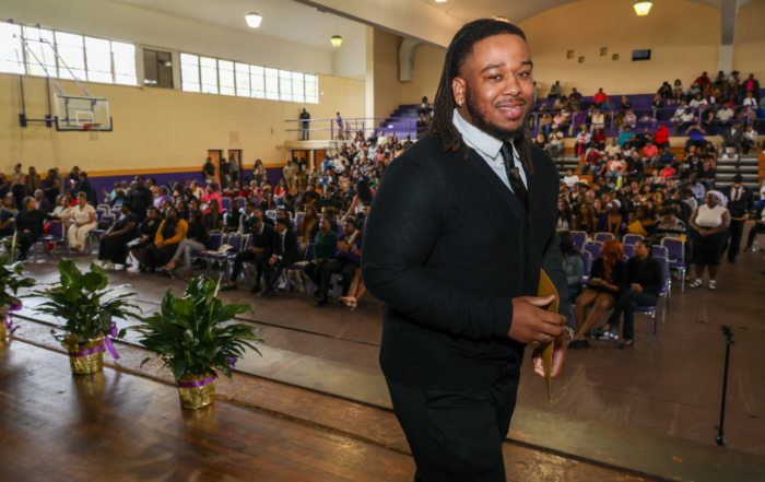 An Alcorn State University student smiles while walking across the stage at E.E. Simmons Gym during the 87th Honors Convocation, holding a gold certificate, with a packed audience of students and guests seated in the bleachers behind him.