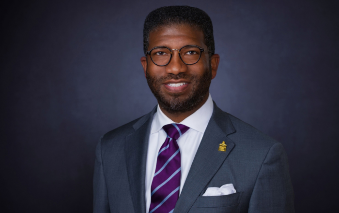 The Rev. C. Edward "CJ" Rhodes II, director of religious and spiritual life at Alcorn State University, poses for a professional headshot wearing a gray suit, purple striped tie and white pocket square against a dark gray background.