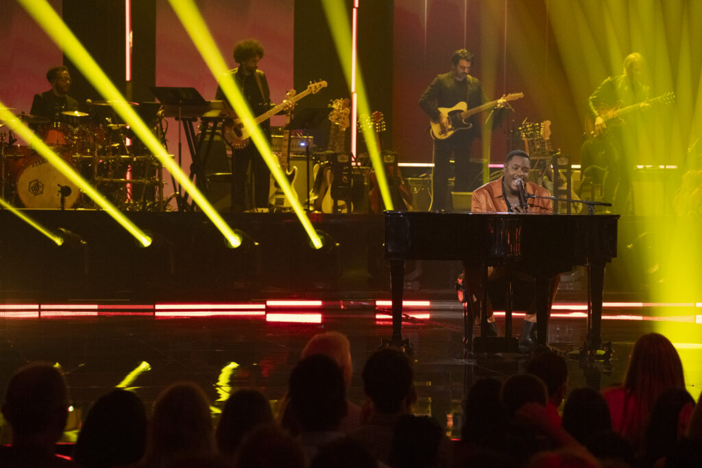 Alcorn State University alumnus Daniel Stallworth performs on the "American Idol" stage, singing into a microphone in a rust-orange leather jacket against a backdrop of yellow stage lighting and live band musicians.