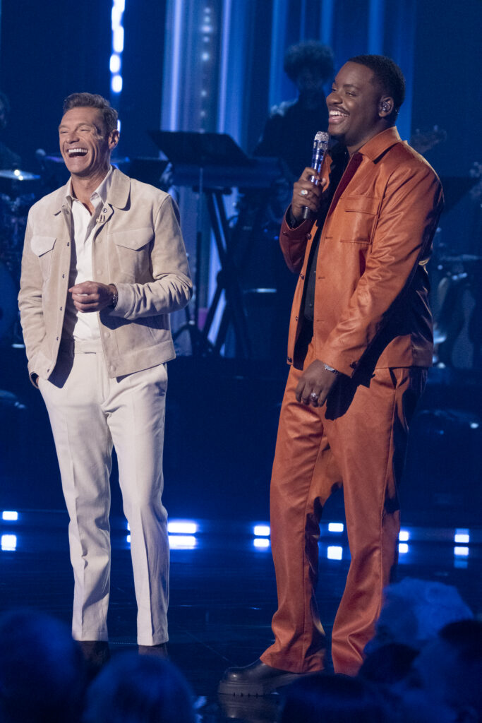 Alcorn State University alumnus Daniel Stallworth stands on the "American Idol" stage holding a microphone alongside host Ryan Seacrest, both laughing, with blue stage lighting in the background.
