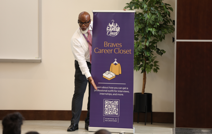 Michael Harper dressed in white button down and black slacks on stage pointing at MyCareerCloset banner, presenting.