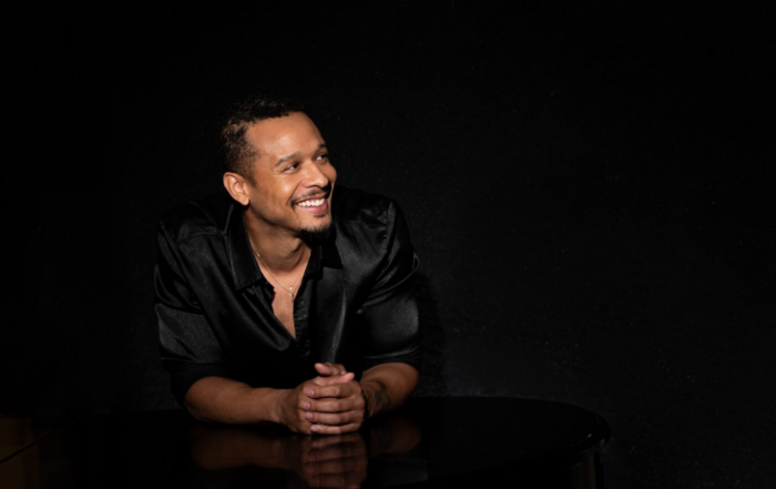 Kortland Whalum, Memphis-based singer-songwriter and headliner of the 44th Annual Alcorn State University Jazz Festival, smiles warmly while seated at a piano, wearing a black satin shirt against a dark studio background.