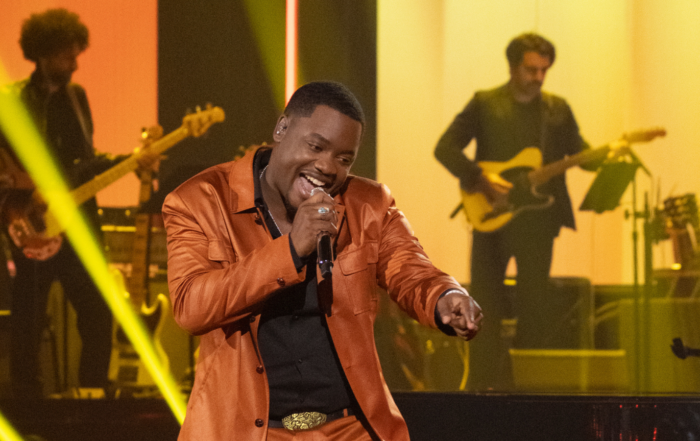 Alcorn State University alumnus Daniel Stallworth performs on the "American Idol" stage, singing into a microphone in a rust-orange leather jacket against a backdrop of yellow stage lighting and live band musicians.