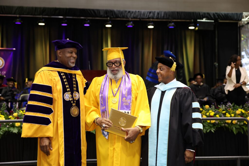 Alcorn State University President Tracy M. Cook and Dr. Marcus Ward, senior vice president for institutional advancement, share a joyful moment on stage with Golden Class of 1976 graduate Andre Young, who holds his diploma and wears a gold cap and gown with a purple Alcorn State Class of 1976 stole during the 2026 Spring Commencement ceremony in Lorman, Mississippi.