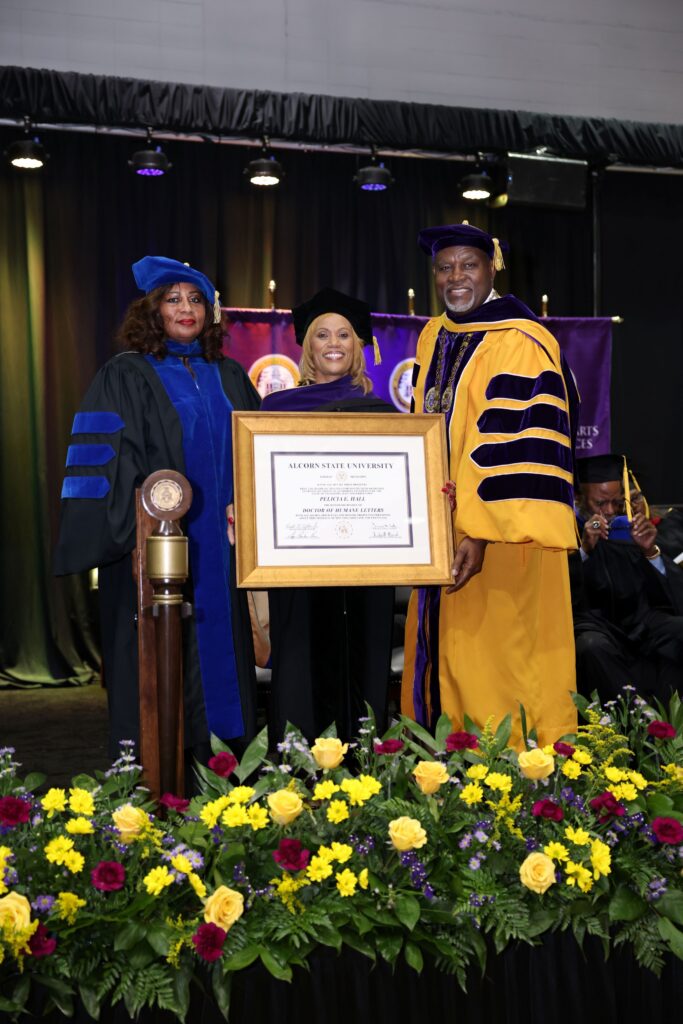 Pelicia Hall receives an honorary Doctor of Humane Letters degree from Alcorn State University during the 2026 Spring Commencement ceremony. Hall stands at center holding the framed degree certificate, flanked by two university officials in academic regalia, with yellow and red floral arrangements in the foreground