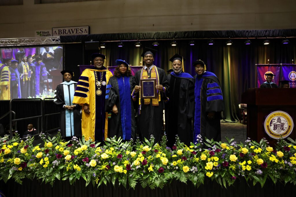 Nigel Caesar, center, holds his Student Award of Excellence plaque on stage during Alcorn State University's 2026 Spring Commencement ceremony, flanked by, from left, Dr. Marcus Ward, President Tracy M. Cook, Provost Joyce Buckner Brown, ASU Foundation Board Chairman Dennis Thomas, and School of Business Dean Antwon Woods, with yellow and red floral arrangements in the foreground and the Alcorn State University seal visible at the podium.