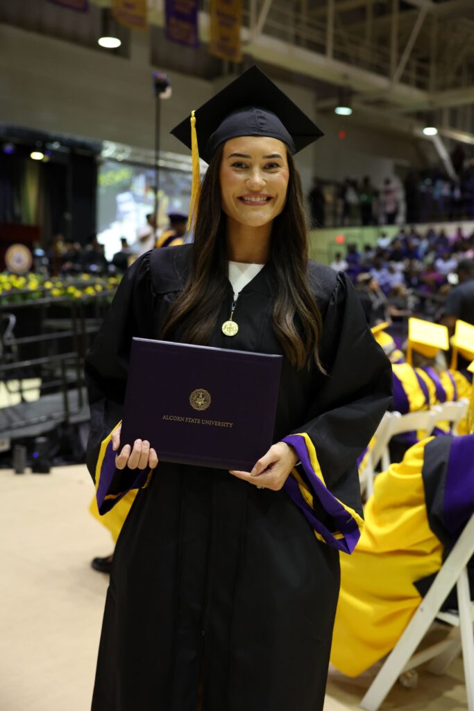 An Alcorn State University graduate smiles while holding her diploma cover during the 2026 Spring Commencement ceremony in the Davey L. Whitney HPER Complex in Lorman, Mississippi. She wears a black cap and gown with purple and gold trim and an Alcorn State University medallion.