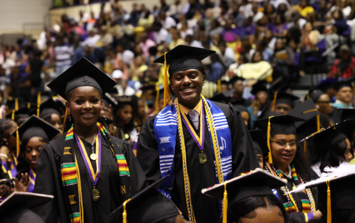 Alcorn State University graduates smile during the 2026 Spring Commencement ceremony in the Davey L. Whitney HPER Complex in Lorman, Mississippi. Graduates wear black caps and gowns adorned with purple and gold honor cords and kente cloth stoles as families fill the stands behind them.