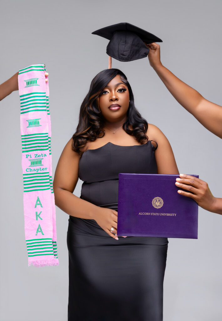 Shantwana Patterson holds an Alcorn State University diploma cover while hands extend a graduation cap and an Alpha Kappa Alpha Pi Zeta Chapter kente stole around her in a graduation portrait.