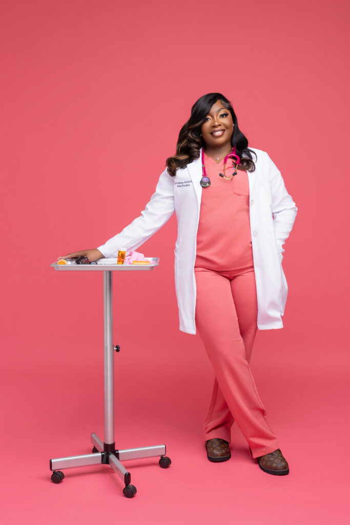 Shantwana Patterson, MSN, FNP, stands confidently in a professional photo shoot setting against a solid pink background. She is wearing coral scrubs, a white lab coat with her name and title visible on the chest pocket, and a pink stethoscope around her neck. She leans with one arm resting on a silver medical tray stand that holds prescription bottles and medical supplies. She is smiling warmly at the camera. Her name tag reads "S. Patterson, MSN/FNP, Nurse Practitioner."