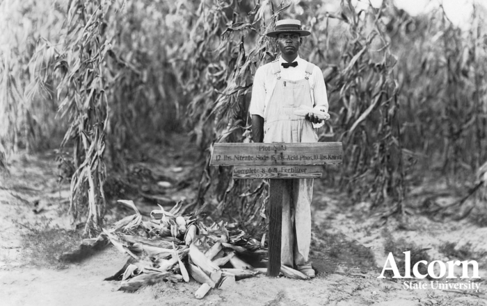 A historic black-and-white photograph of a man in overalls and a straw hat standing beside a wooden fertilizer plot sign in a cornfield, representing Alcorn State University's agricultural land-grant heritage. The Alcorn State University logo appears in the lower right corner.
