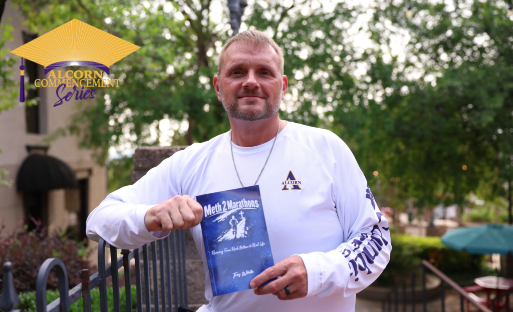 Alcorn State University graduating senior John "Trey" White holds a copy of his book, Meth 2 Marathons: Running From Rock Bottom to Real Life, outside on campus wearing an Alcorn long-sleeve shirt.