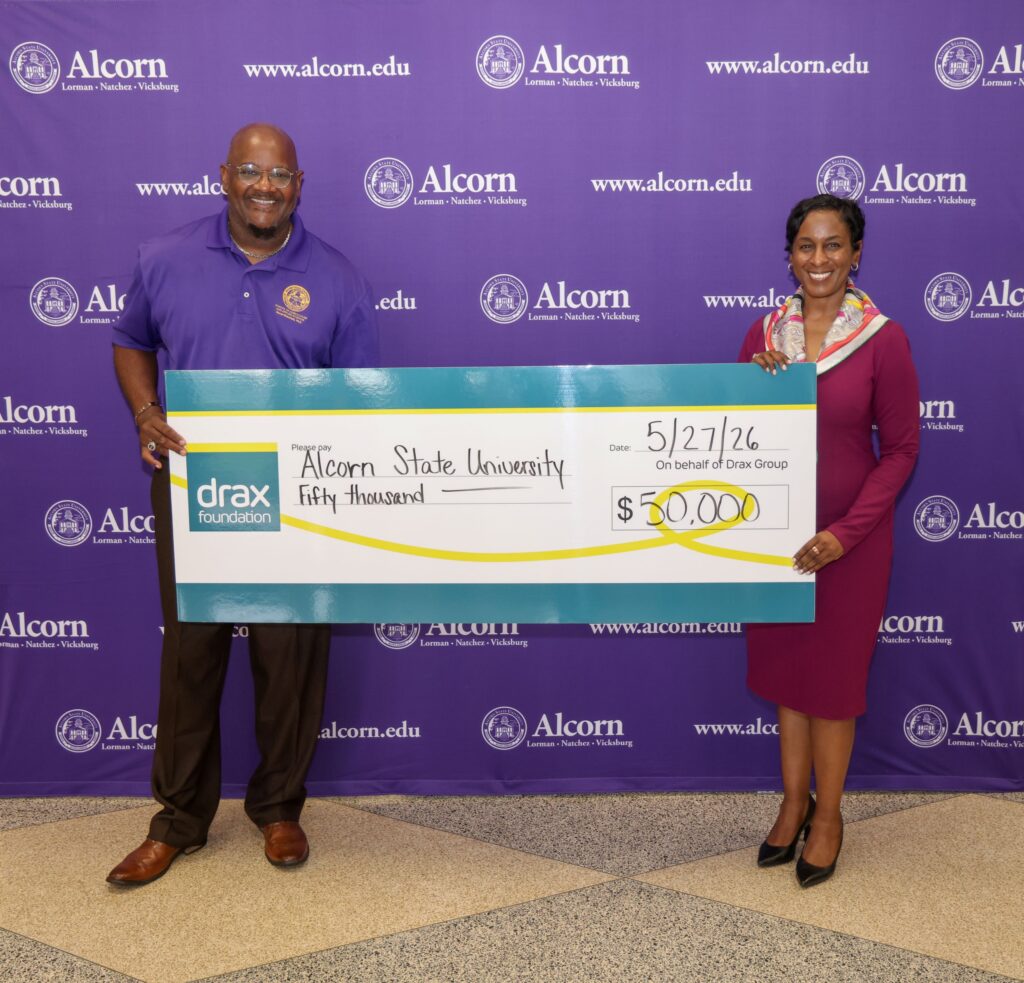 Dr. Dexter B. Wakefield I, dean of Alcorn State University's College of Agriculture and Applied Sciences, wearing a purple Alcorn polo, and Deidra Jackson, senior vice president of corporate affairs at Drax, wearing a magenta dress, smile while holding an oversized Drax Foundation check made out to Alcorn State University for $50,000, dated 5/27/26, in front of a purple Alcorn State University step-and-repeat backdrop.