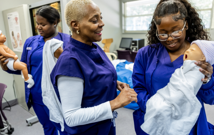 Three Alcorn State University nursing students in purple scrubs practice swaddling and holding infant-simulator manikins in a clinical skills lab, with anatomical charts and simulation equipment in the background.