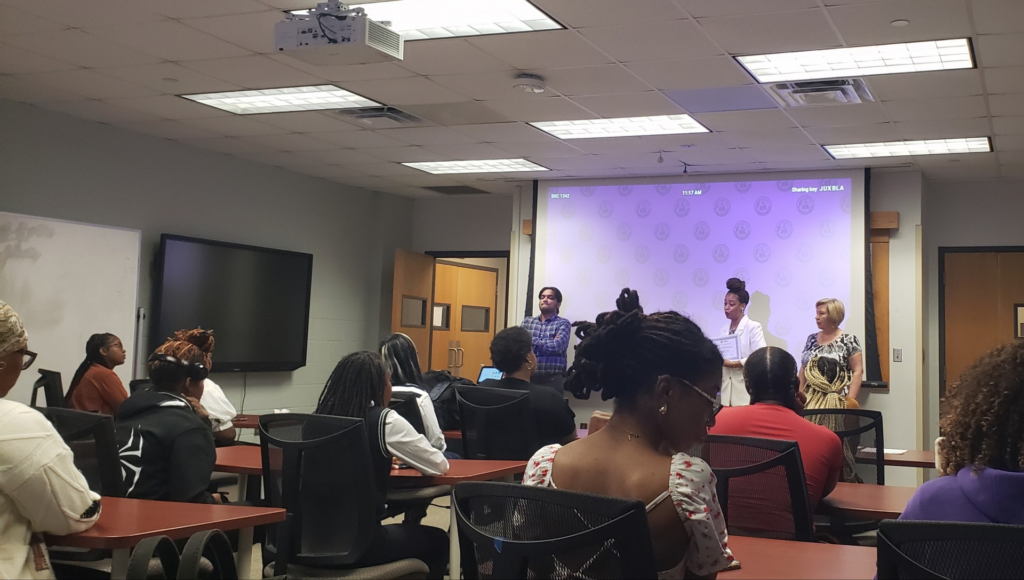 Students seated at tables in a classroom watch a presentation at the front of the room. 