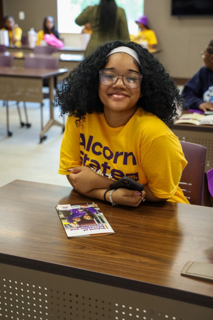 A TMCF SOAR scholar smiles at the camera while seated at a desk during the Meet the Deans and Major Exploration session at Alcorn State University. She wears a gold Alcorn State t-shirt and clear-framed glasses, with an Alcorn State School of Education and Psychology program brochure on the desk in front of her. Other participants are visible in the background of the classroom setting.