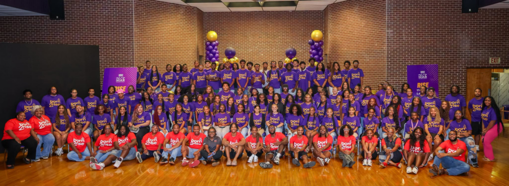More than 100 TMCF SOAR scholars and staff pose for a group photo inside Alcorn State University's gymnasium, wearing purple and red "Born to Rise" t-shirts surrounded by purple and gold balloons.