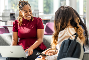 Three women conversing during a career fair-- the two students are facing away from the camera, while the other woman has a laptop in her lap as she speaks to the students.