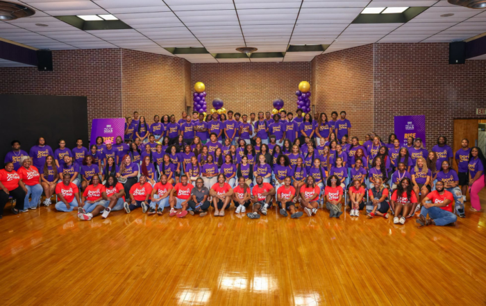 A large group photo of more than 100 students and staff gathered inside a gymnasium at Alcorn State University for the 2026 TMCF SOAR Program. Students wear purple "Born to Rise" t-shirts and staff wear red "Born to Rise" t-shirts. The group is arranged in tiered rows, with purple and gold balloon columns and TMCF SOAR branded banners visible in the background.