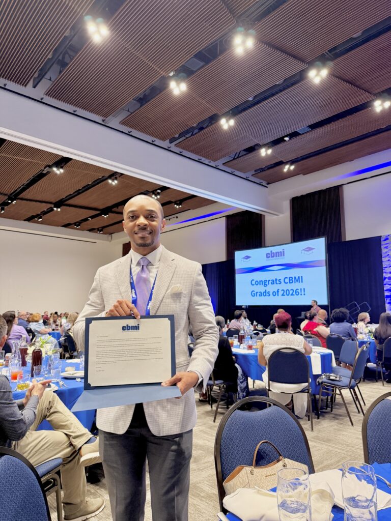 Casey Mock holds his CBMI certificate at a banquet, with a screen reading "Congrats CBMI Grads of 2026!!" on the wall behind him.