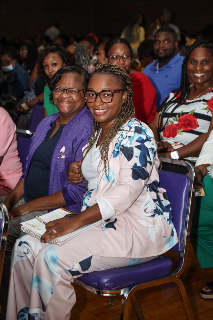Sonia Eley and Kisha Bond smile for the camera while seated in a crowded audience.