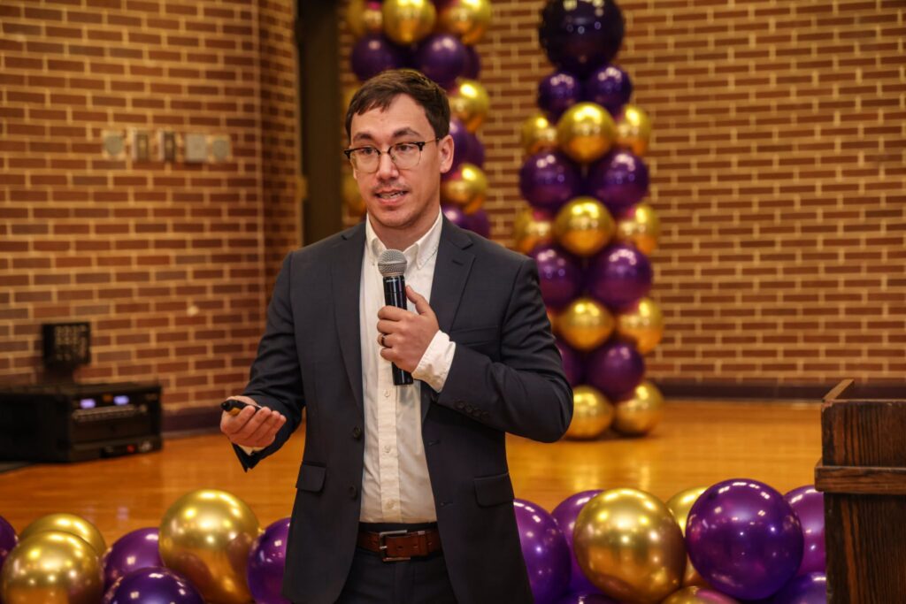 Kyle Kubovcik of EAB Navigate speaks into a handheld microphone while gesturing during a presentation, standing near a podium with purple and gold balloon columns behind him.