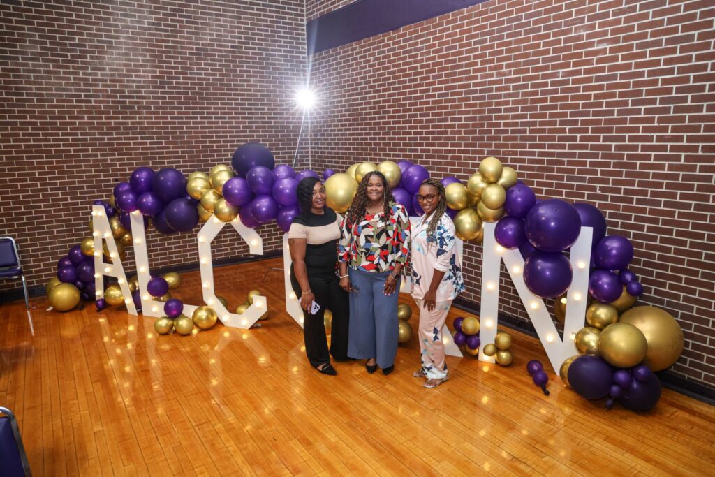Three women stand together and smile for a photo in front of large illuminated marquee letters spelling "ALCORN," framed by a purple and gold balloon garland against a brick wall.