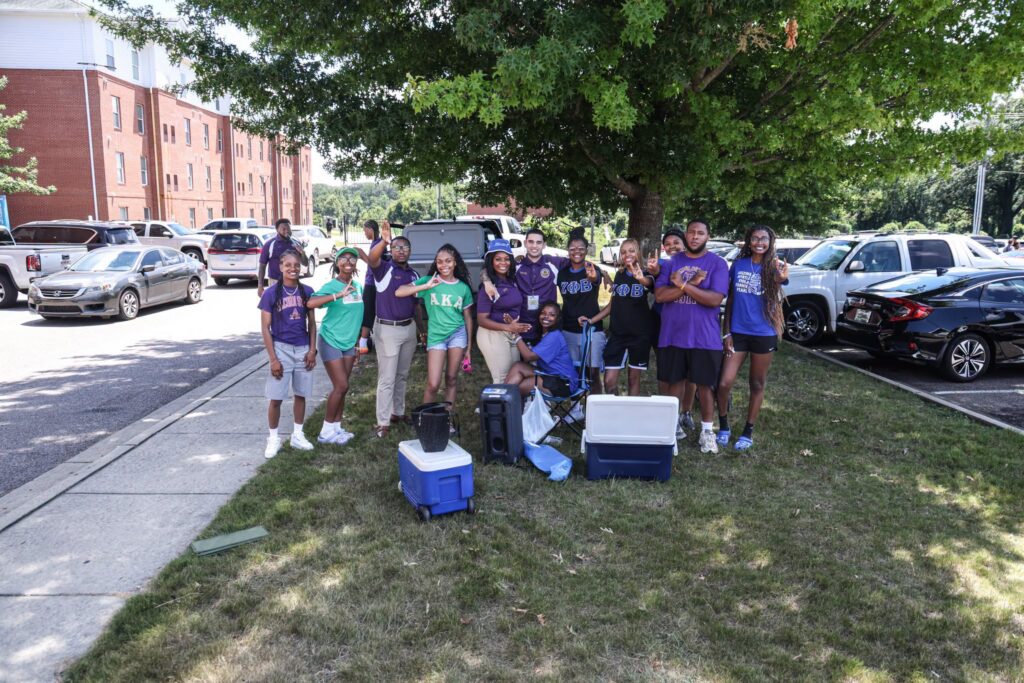 Members of several National Pan-Hellenic Council organizations pose together under a shade tree near a residence hall, with coolers and a fan set up to help during Move-In Day.