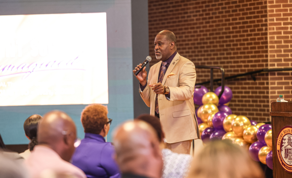 Alcorn State University President Tracy M. Cook, in a tan suit and purple tie, speaks into a handheld microphone during the Faculty and Staff Institute, gesturing toward an audience seated in the foreground, with a "Student Success Reimagined" screen, purple and gold balloons, and an Alcorn State-seal podium behind him.