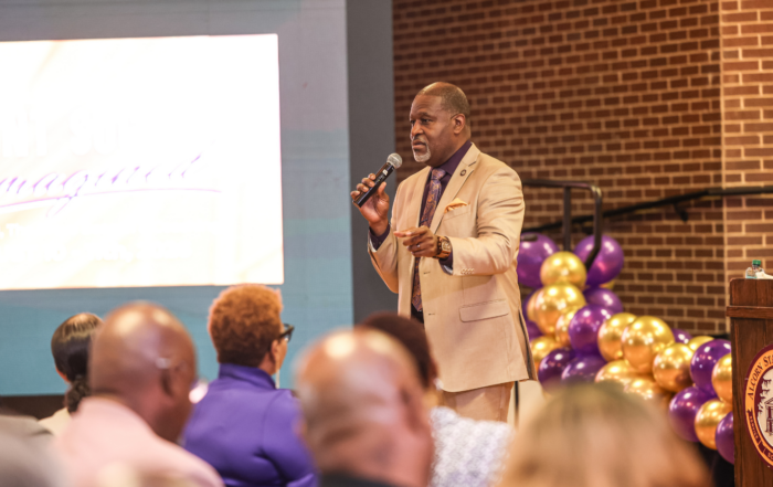 Alcorn State University President Tracy M. Cook, in a tan suit and purple tie, speaks into a handheld microphone during the Faculty and Staff Institute, gesturing toward an audience seated in the foreground, with a "Student Success Reimagined" screen, purple and gold balloons, and an Alcorn State-seal podium behind him.