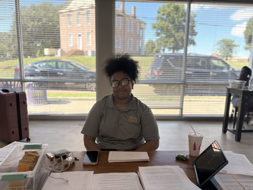 A resident assistant in an Alcorn Department of Residence Life polo sits at a check-in table stacked with housing paperwork during Move-In Day.