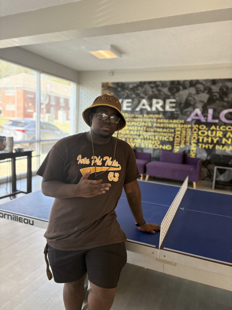 Young man in a brown Iota Phi Theta T-shirt and bucket hat stands by a ping-pong table in an Alcorn residence hall lounge.