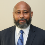 Portrait of Dr. Nolan McMurray, dean of Alcorn State University's College of Arts and Sciences, wearing a dark suit with a white shirt and blue striped tie, against a tan background.