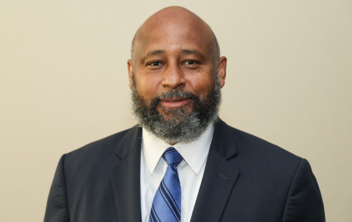 Portrait of Dr. Nolan McMurray, dean of Alcorn State University's College of Arts and Sciences, wearing a dark suit with a white shirt and blue striped tie, against a tan background.