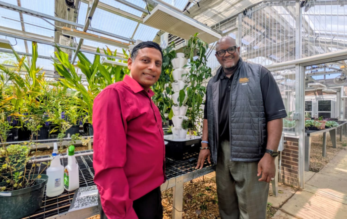 Dr. Emran Ali, left, in a red shirt, and Dr. Dexter Wakefield, right, in a dark vest with an Alcorn logo, smile as they stand in a greenhouse beside a white vertical hydroponic tower of planting pockets. Potted plants fill the benches around them.