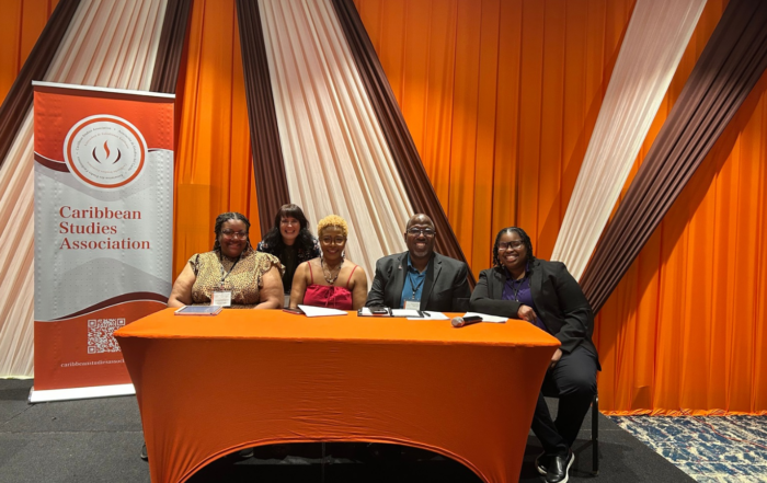 From left, Precious Cooper, Donna Hope, Byron Johnson, Latuesday Guy and Heather Bailey smile behind an orange-covered table beside a Caribbean Studies Association banner. Four are seated, and one stands behind them.