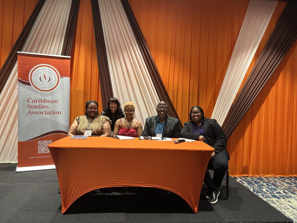   From left, Precious Cooper, Donna Hope, Byron Johnson, Latuesday Guy and Heather Bailey smile behind an orange-covered table beside a Caribbean Studies Association banner. Four are seated, and one stands behind them.