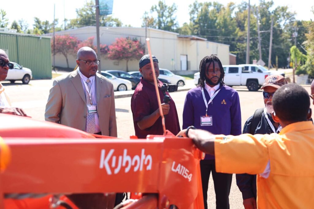 Attendees wearing conference lanyards stand in a parking lot and listen to a presenter in a yellow shirt, seen from behind, who rests a hand on the loader of an orange Kubota tractor. One man holds a camera on a handheld grip. Buildings and trees are behind the group.