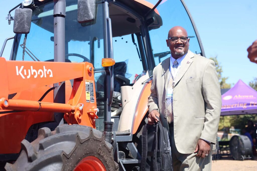 Attendees wearing conference lanyards stand in a parking lot and listen to a presenter in a yellow shirt, seen from behind, who rests a hand on the loader of an orange Kubota tractor. One man holds a camera on a handheld grip. Buildings and trees are behind the group.
