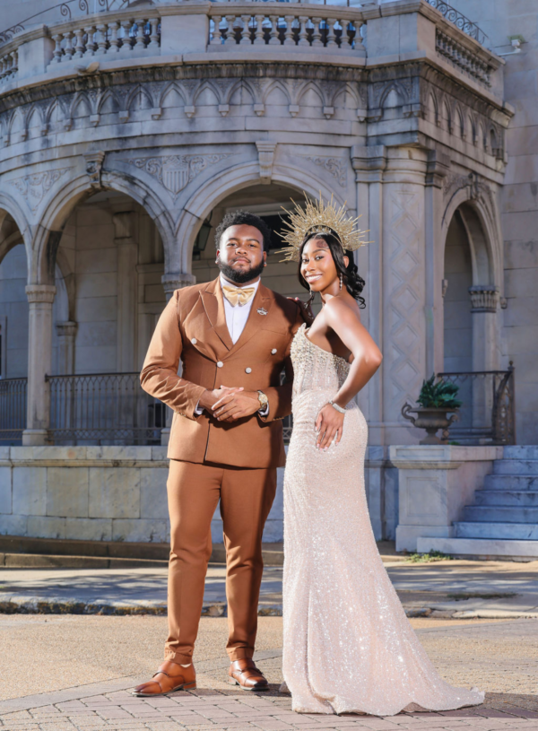 Jerrell Ervin, Mister Alcorn, and Erin Nichols, the 100th Miss Alcorn, pose in formal coronation attire in front of an ornate historic building with classical architecture. Ervin wears a tan double-breasted suit with a white dress shirt and pearl accessories. Nichols wears a champagne-colored strapless sequined gown with a gold spiked crown headpiece. Behind them is an elegant stone facade featuring arched doorways, decorative columns, and carved detailing characteristic of neoclassical architecture.